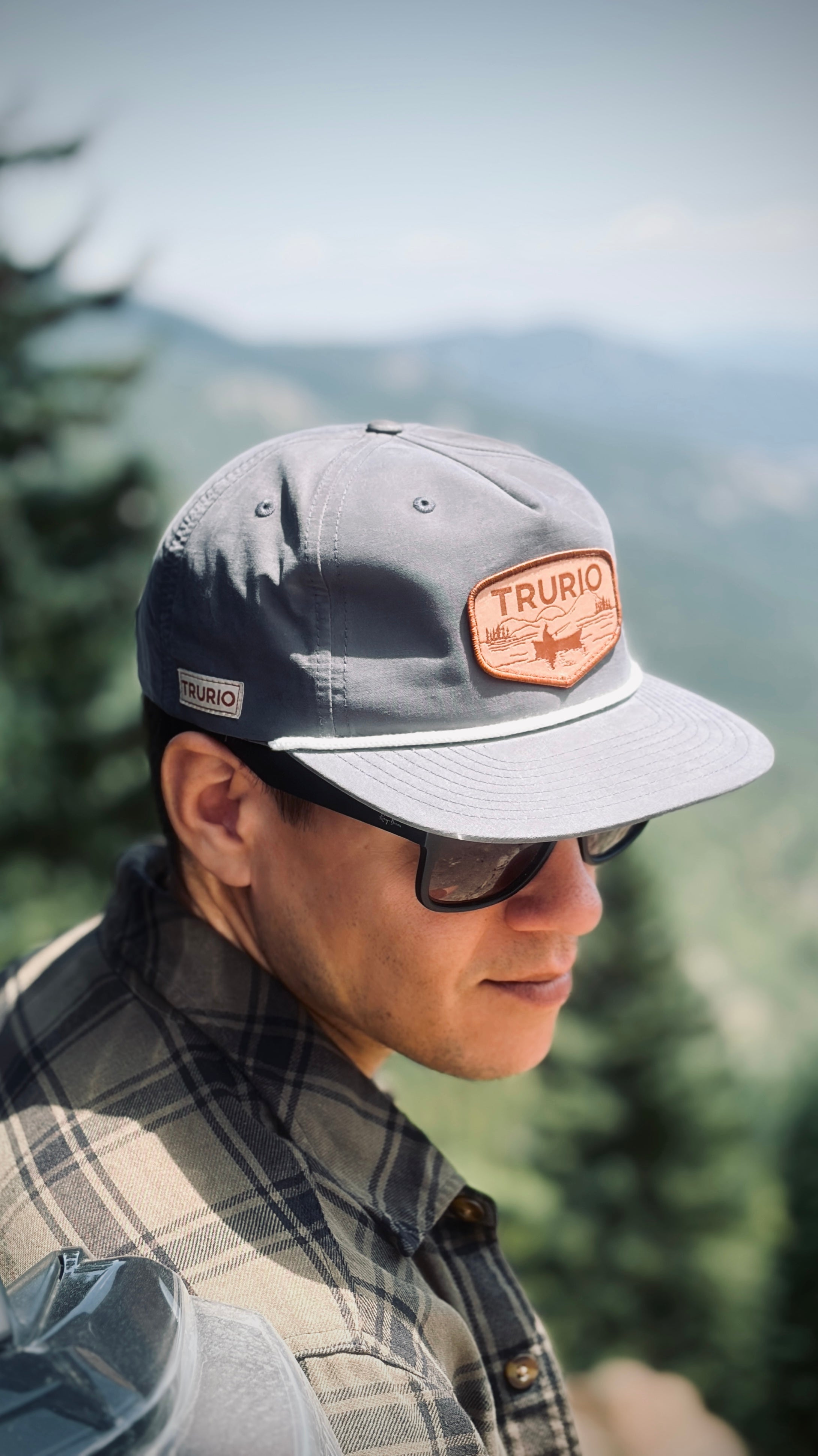 A man wearing a gray Trurio 5-star cap with a brown leather patch, black sunglasses, and a plaid shirt, photographed outdoors against a blurred mountain background.